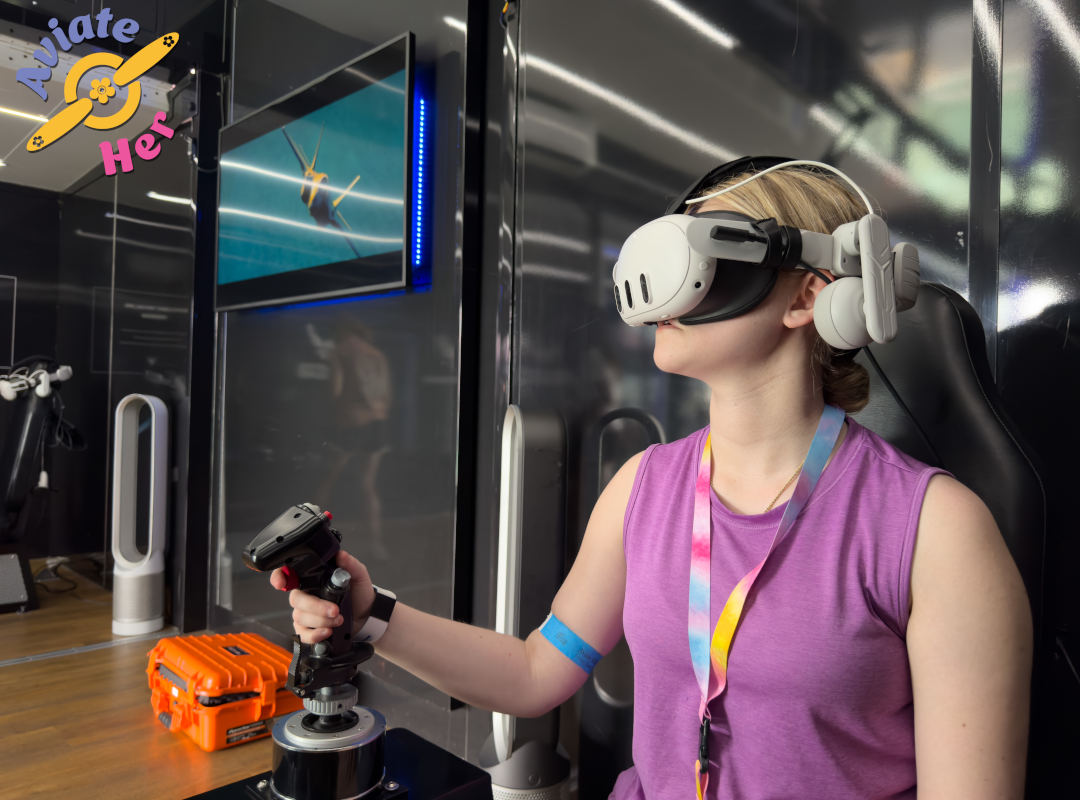 Aviate Her A young woman participates in the “Aviate HER” event at the Dayton Air Show, wearing a virtual reality headset and holding a flight simulator joystick while seated in a booth. A monitor in front of her shows an aircraft in flight, and the Aviate HER logo with a propeller and flower design is displayed on the wall. She wears a tie-dye lanyard and purple shirt, highlighting the event’s focus on inspiring women in aviation through interactive technology.