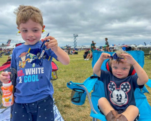 Two young children at the Dayton Air Show hold toy Blue Angels jets while sitting on lawn chairs in the viewing area. Wearing ear protection and patriotic clothing, they enjoy the family-friendly aviation event surrounded by other spectators under a cloudy sky.
