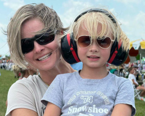 A smiling woman and young child enjoy the Dayton Air Show, both wearing sunglasses while the child also wears protective ear muffs for the loud aircraft. They sit among the crowd on a sunny day, highlighting the family-friendly atmosphere of the aviation event.
