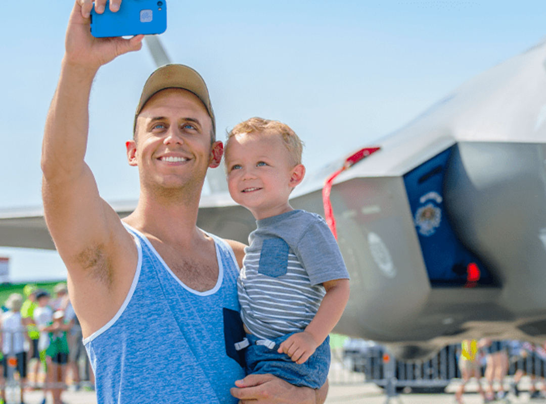 Early Access A smiling man in a blue tank top holds a young child while taking a selfie in front of a modern fighter jet on display at an airshow, capturing the excitement of early access admission.