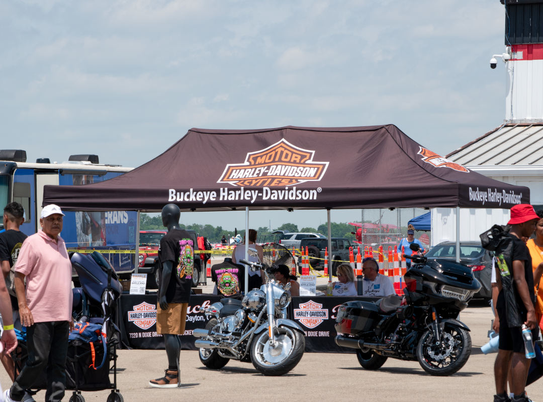 Exhibitor A Buckeye Harley-Davidson exhibitor display at an outdoor air show features a branded canopy tent with the Harley-Davidson Motor Cycles logo, two showcased motorcycles parked beneath the tent, and a mannequin wearing a colorful Harley-Davidson T-shirt. People walk by the booth, including attendees carrying bags and drinks, while event staff sit at a table under the tent with raffle signs. The setting is a sunny day with vehicles, orange traffic cones, and other vendor tents visible in the background.