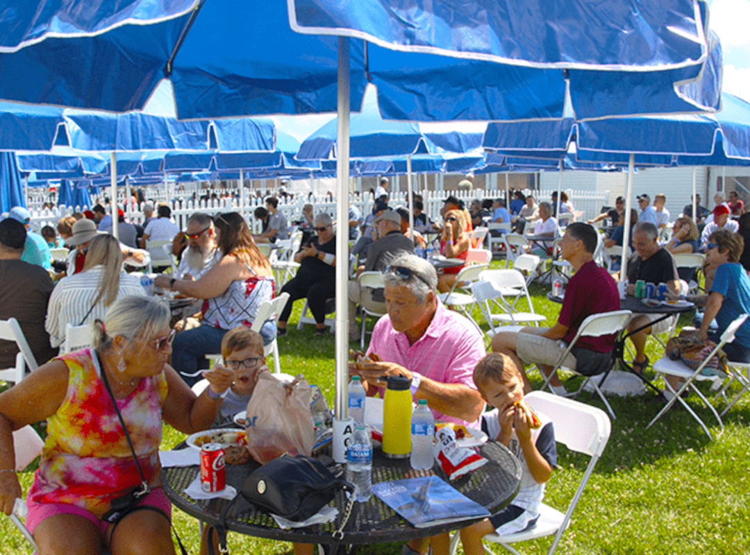 Private Chalets Families and guests enjoy food and drinks under shaded blue umbrellas in a private chalet area at an airshow, creating a relaxed VIP dining experience with reserved seating.