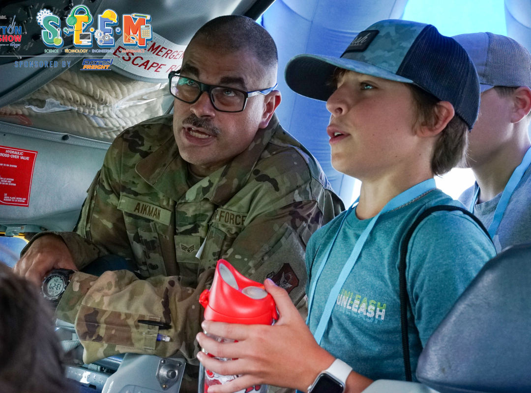 STEM display A U.S. Air Force service member in camouflage uniform explains aircraft systems to a young attendee inside a STEM display at the Dayton Air Show. The boy listens attentively while holding a water bottle, wearing a blue “Unleash” T-shirt and event lanyard. The STEM program logo, promoting science, technology, engineering, and math, is visible in the corner, highlighting the event’s educational aviation outreach.