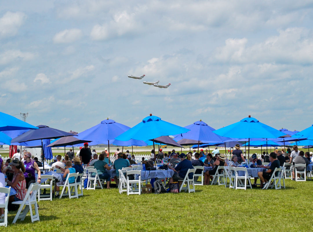 Chairman's Club Guests relax under blue sun umbrellas in the Chairman’s Club at the Dayton Air Show, a premium seating area offering shaded tables, catered dining, and front-row views of aerial performances. In the background, a formation of three aircraft soars across the sky, giving attendees an exclusive, comfortable vantage point to enjoy the aviation spectacle.