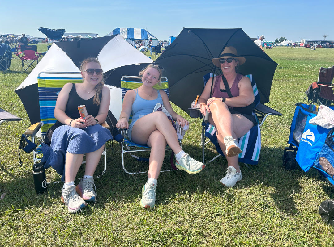 General Admission Three women relax in the General Admission area at the Dayton Air Show, seated in lawn chairs with umbrellas for shade on a sunny day. They enjoy drinks and snacks while watching the aviation performances from the open grassy field, reflecting the casual, family-friendly atmosphere available to all attendees.