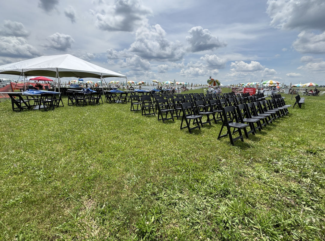 P-51 Club The P-51 Club seating area at the Dayton Air Show features neatly arranged black chairs and shaded tables under a large white tent, offering guests a comfortable and organized viewing space close to the runway. Surrounded by open green grass and colorful umbrellas in the distance, the section provides a relaxed yet premium atmosphere for enjoying the aviation performances.