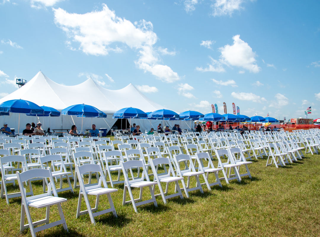 Skyview Lounge The Skyview Lounge at the Dayton Air Show features rows of white chairs on the grass with shaded tables under blue umbrellas, positioned in front of a large white hospitality tent. Guests enjoy a comfortable seating arrangement with clear views of the runway, combining relaxation, shade, and an elevated experience for watching the aviation performances.
