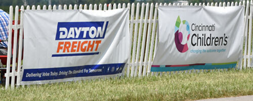 dassigns Images of sponsor banners in the distance on a fence. They are approximately two to eight feet wide and 2 feet tall. Various colors.