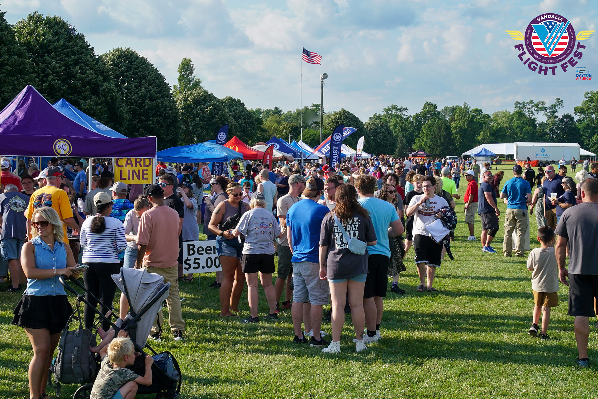 2025_DAS-122 Children and families line up at a colorful booth during Flight Fest in Vandalia, Ohio. Under a bright teal tent with the words “Know. Love. Serve. Simplify.” volunteers hand out materials and provide face painting, while inflatable attractions and festival activities are visible in the background. The scene highlights the family-friendly, community celebration connected to the aviation-themed event.