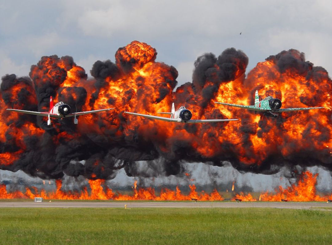 Three vintage World War II–era aircraft perform a low-level formation flyover in front of a massive wall of orange fire and black smoke during the “Tora! Tora! Tora!” airshow reenactment. The dramatic explosion effect simulates a Pearl Harbor battle scene, with the planes flying just above the runway as flames and smoke billow behind them against a cloudy sky