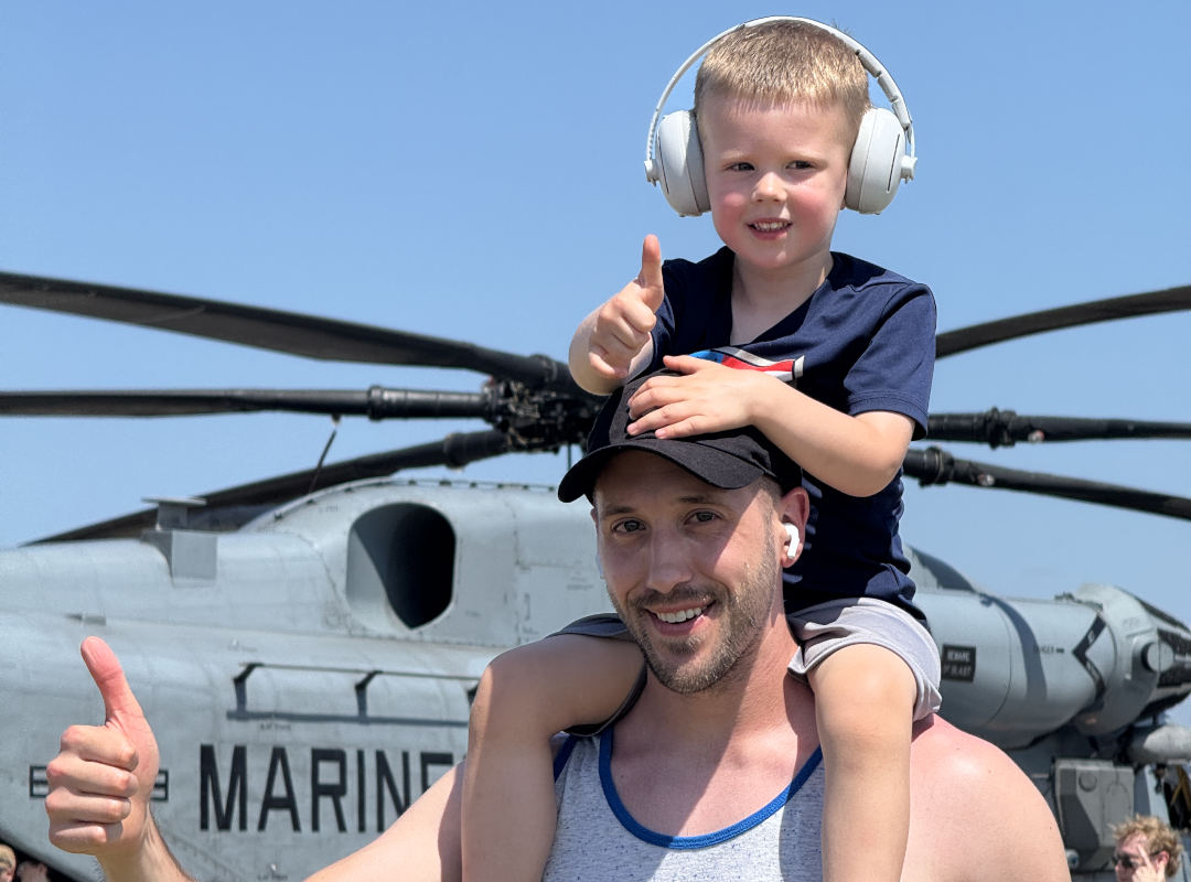 cpdas-2026-schedule-page A smiling man in a blue tank top holds a young child while giving a thumbs up in front of a modern helicopter on display at an air show.