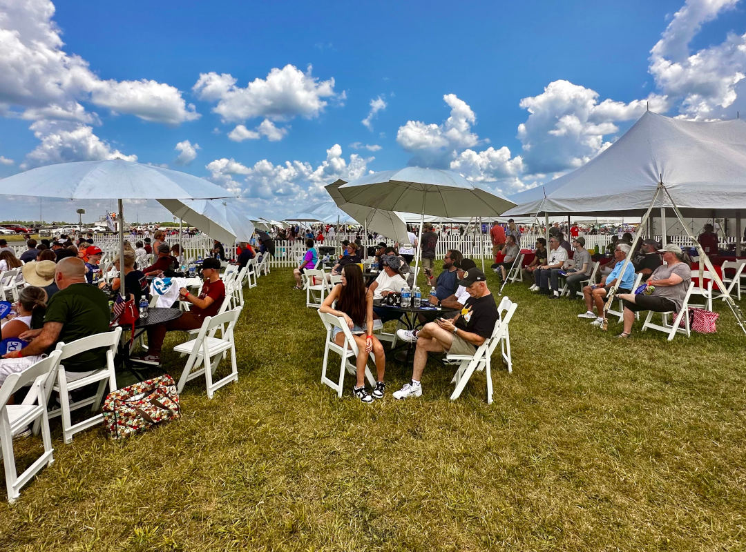 Aviators Alley Guests seated at white folding tables under umbrellas and tents at the Aviators Alley VIP area of an outdoor air show. People are relaxing, eating, and watching the event on a sunny day with blue skies and scattered clouds.