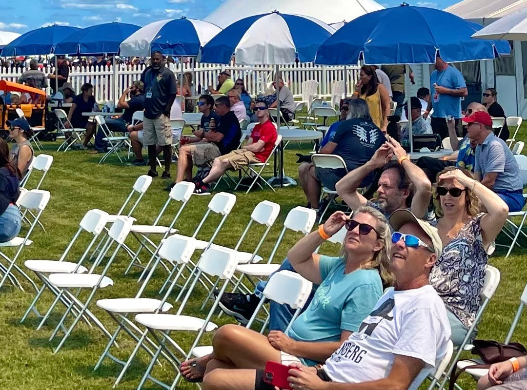 Blue Sky Chalet Spectators sitting on white folding chairs and under blue umbrellas in the Blue Sky Chalet section of an outdoor air show. Families and friends look up at the sky, some shading their eyes with sunglasses, while enjoying the sunny weather