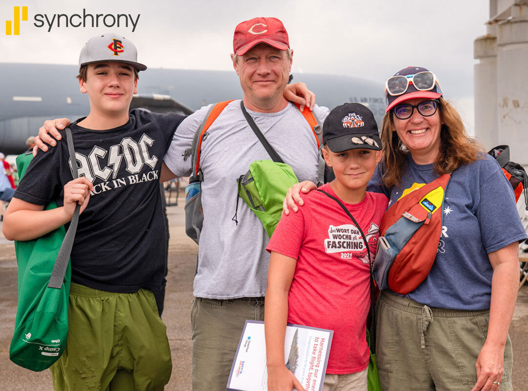 Family 4-pack Smiling family of four posing together at an outdoor air show, carrying backpacks and wearing casual clothes and hats. The parents and two children stand arm in arm, ready to enjoy the event.