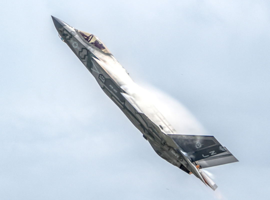 U.S. Navy F-35C Lightning II stealth fighter jet climbing sharply against a pale sky, showing vapor condensation over the wings during a high-performance maneuver.