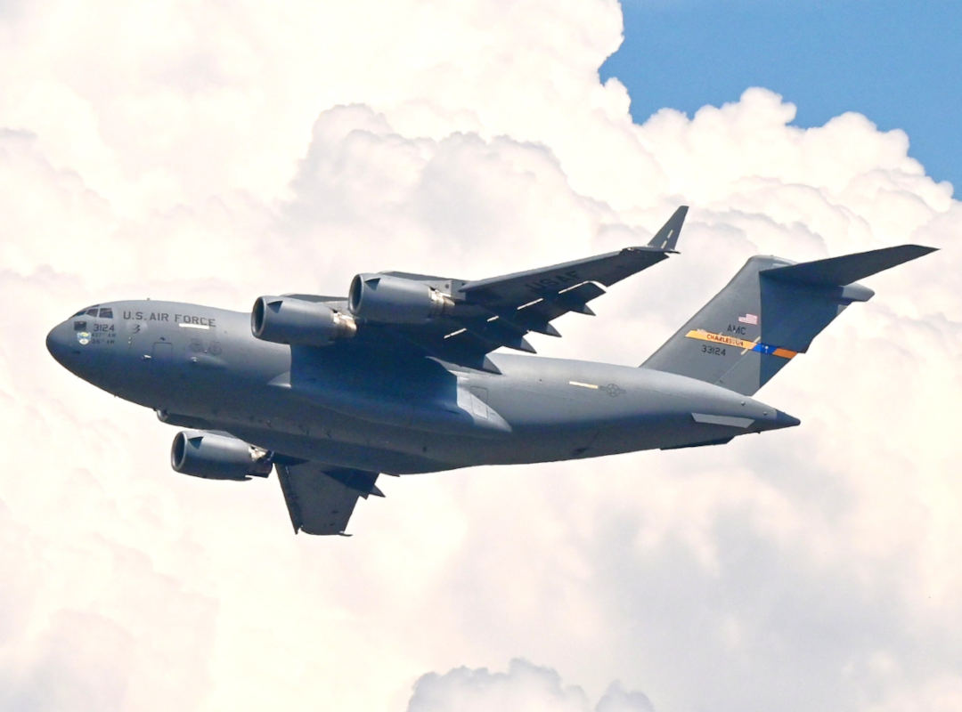 U.S. Air Force C-17 Globemaster III military transport aircraft flying against a partially cloudy sky, showing its four engines and high-mounted wings.