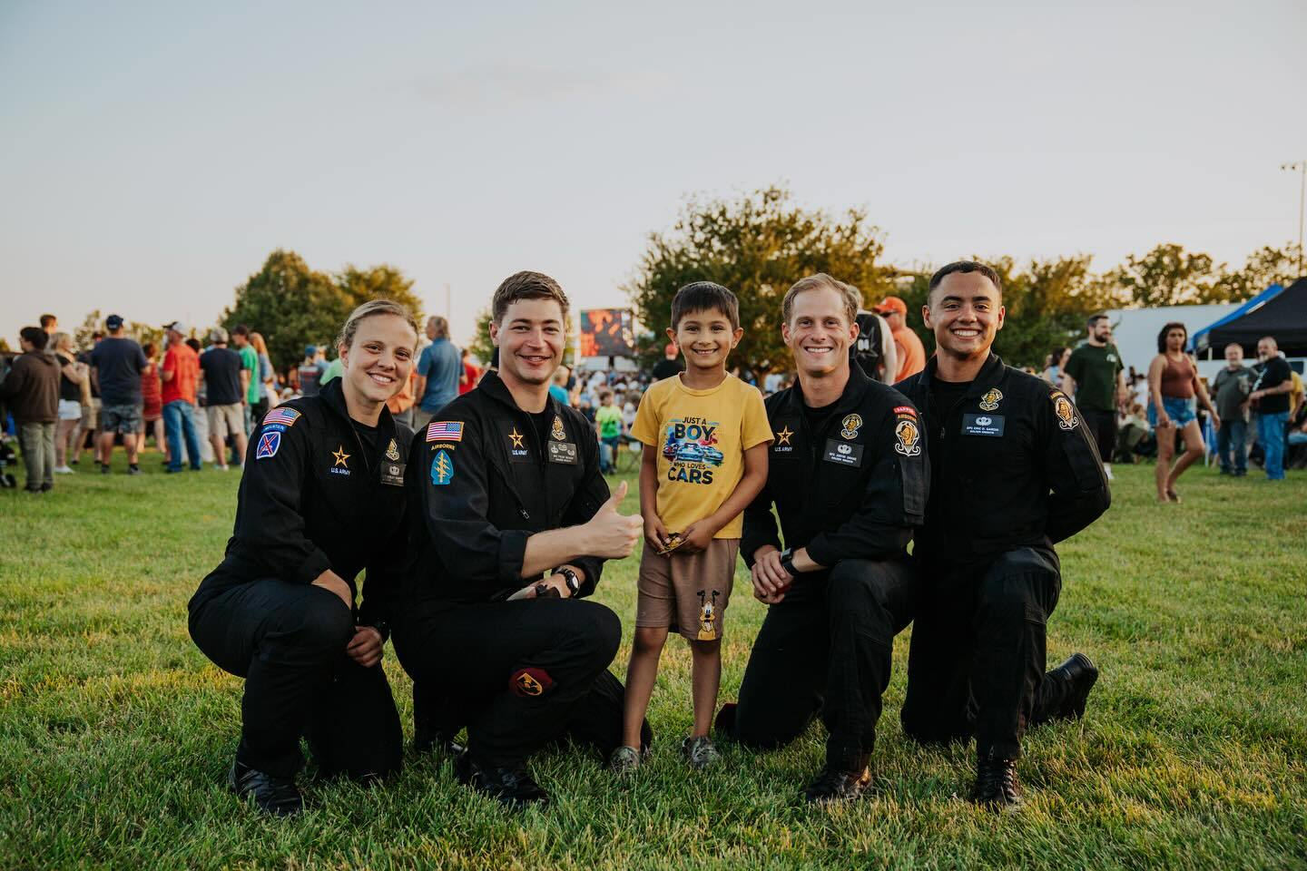 Four U.S. Army soldiers in black flight uniforms kneel on the grass posing with a smiling child during the Dayton Air Show Vandalia Flight Fest, with event attendees and a large outdoor screen visible in the background.