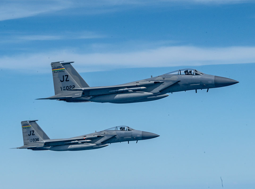 Two U.S. Air Force F-15 fighter jets flying in close formation against a blue sky.