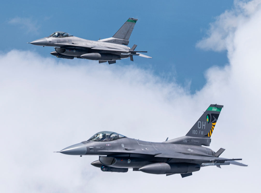 Two U.S. Air Force F-16 fighter jets flying in formation against a blue sky with scattered clouds.