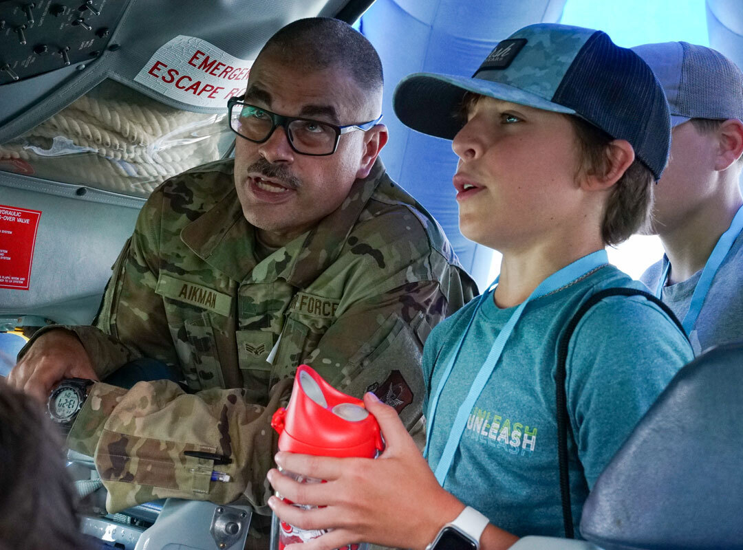 A U.S. Air Force service member in camouflage uniform explains aircraft systems to a young attendee inside a STEM display at the Dayton Air Show. The boy listens attentively while holding a water bottle, wearing a blue “Unleash” T-shirt and event lanyard. The STEM program logo, promoting science, technology, engineering, and math, is visible in the corner, highlighting the event’s educational aviation outreach.