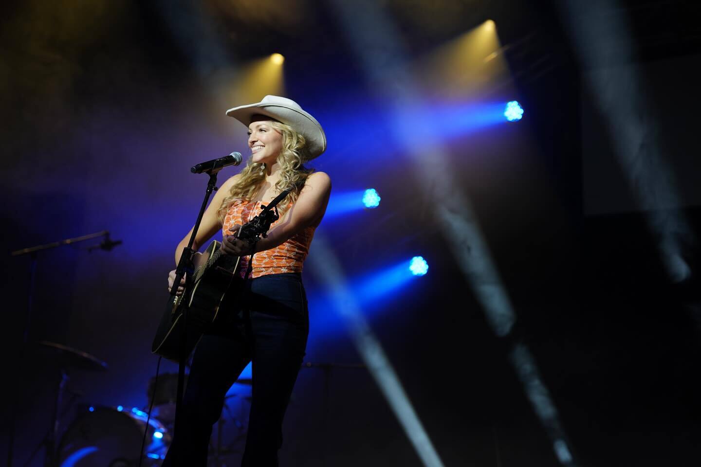 Alexis Gomez, American Idol Top Eight finalist and Dayton native, performs on stage with a guitar and microphone under blue and yellow stage lighting.