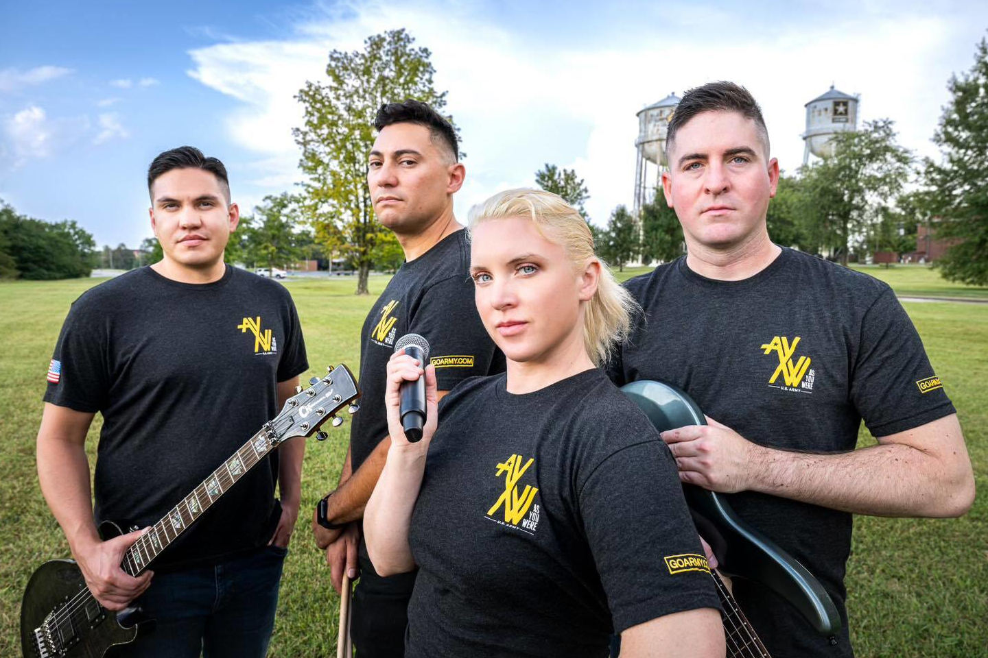 Members of the U.S. Army Corps Band stand together outdoors holding guitars and a microphone while wearing black Army-branded shirts, with water towers and trees visible behind them.