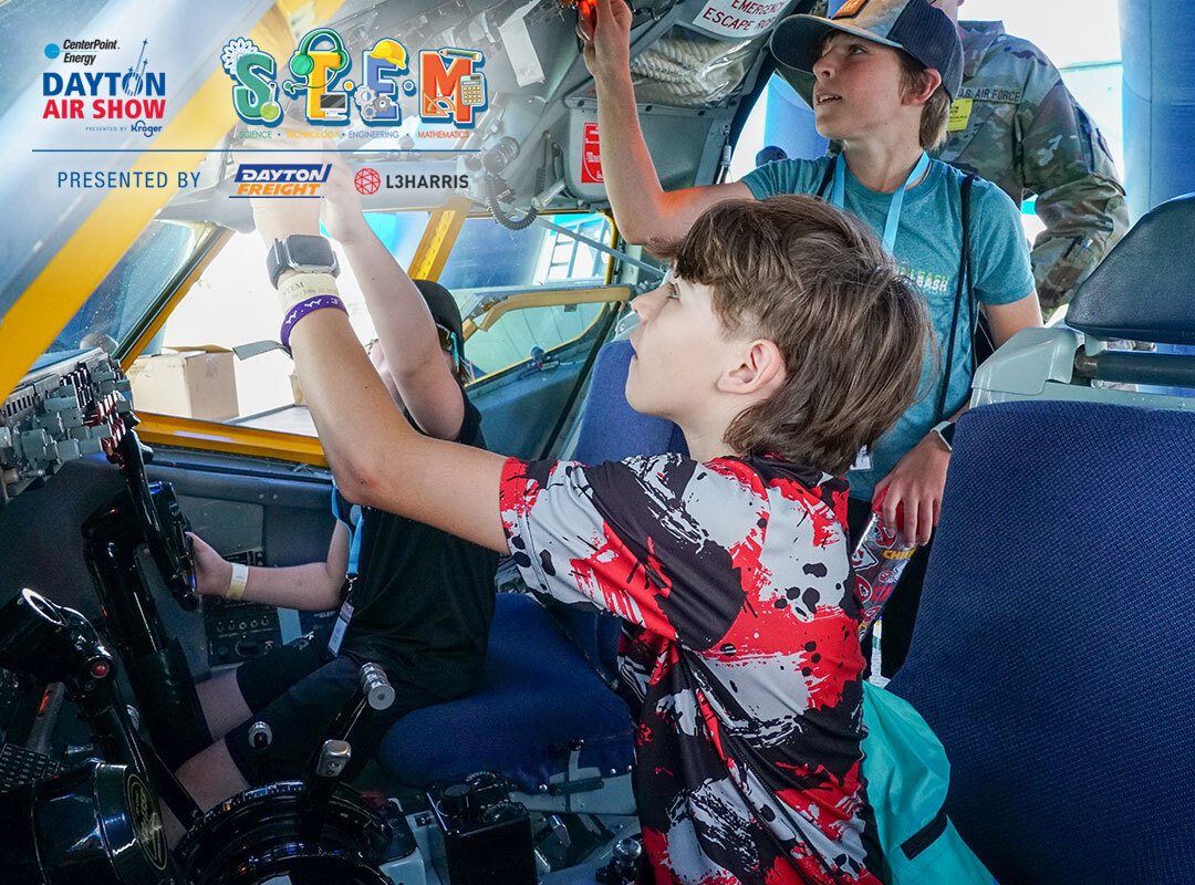Children participate in a hands-on STEM activity inside an aircraft cockpit at the Dayton Air Show, interacting with controls and instrumentation while exploring aviation technology; event branding and sponsor logos including Dayton Freight and L3Harris are visible.
