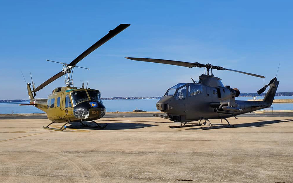 A UH-1 Huey and an AH-1F Cobra helicopter parked side by side on the tarmac near the water, offered as ride experiences at the 2026 Dayton Air Show.
