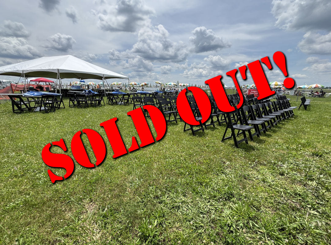The P-51 Club seating area at the Dayton Air Show features neatly arranged black chairs and shaded tables under a large white tent, offering guests a comfortable and organized viewing space close to the runway. Surrounded by open green grass and colorful umbrellas in the distance, the section provides a relaxed yet premium atmosphere for enjoying the aviation performances. This area is sold out for this year's show. 