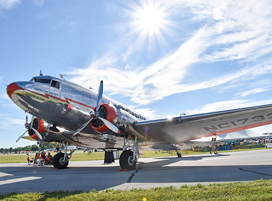 Douglas DC-3 aircraft parked on ramp with sunlight and ground crew nearby