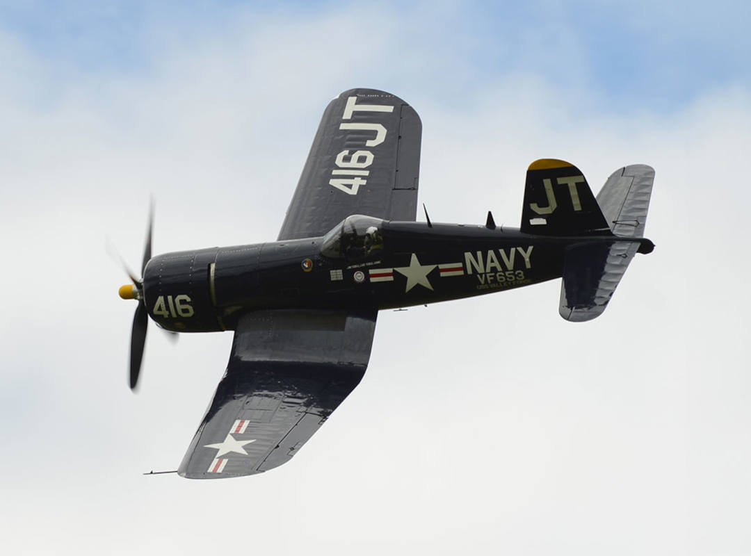 Side profile of a dark blue Vought F4U Corsair warbird flying against an overcast sky with landing gear retracted and propeller in motion.