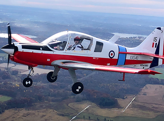 Scottish Aviation Bulldog in flight over rural landscape with pilot visible