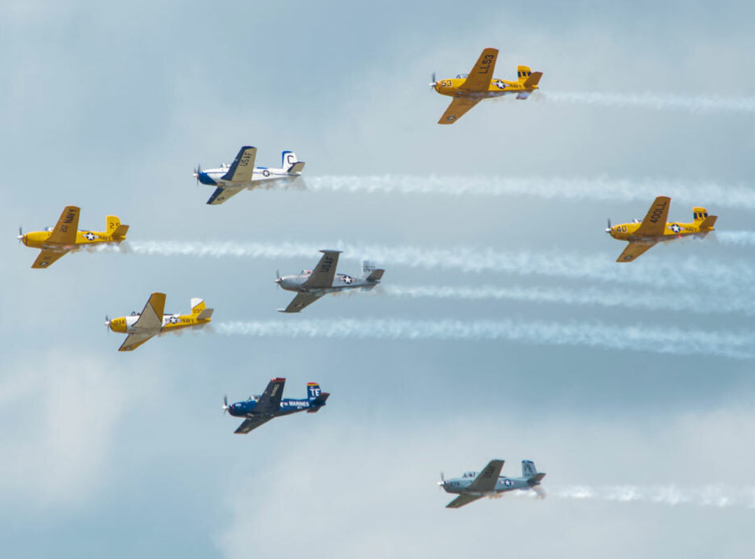 Multiple T-34 Association aircraft flying in formation with smoke trails during an air show performance against a cloudy sky, featuring yellow, silver, blue, and gray military-style paint schemes.
