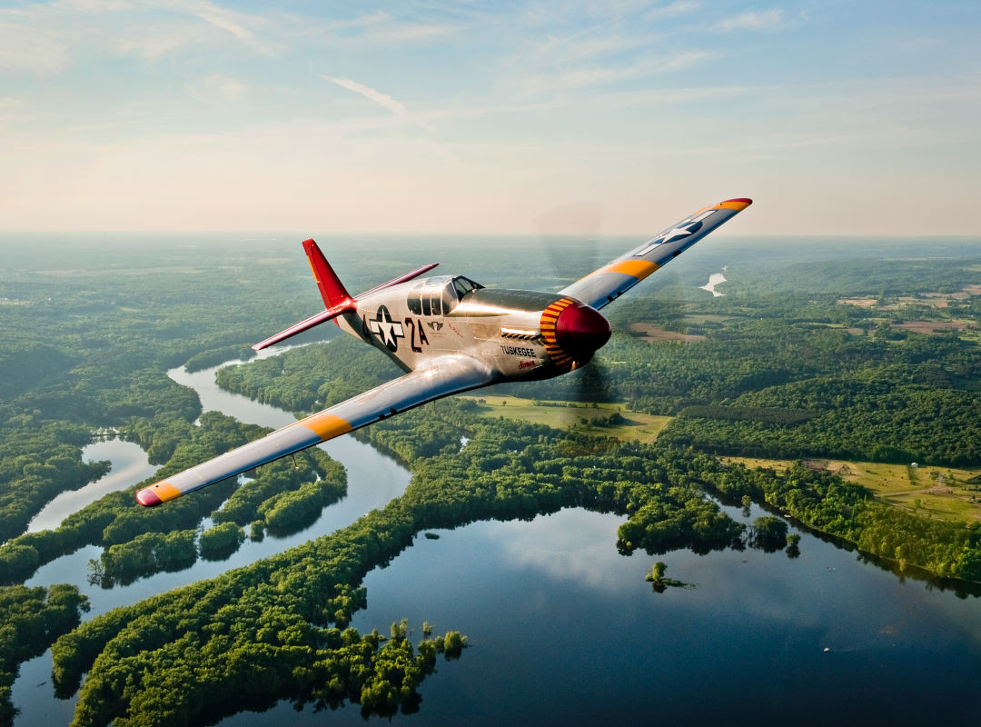 P-51 Mustang “By Request” flying above a winding river and forested landscape during daylight, viewed from above with polished silver fuselage, red tail, and yellow wing accents visible in flight.