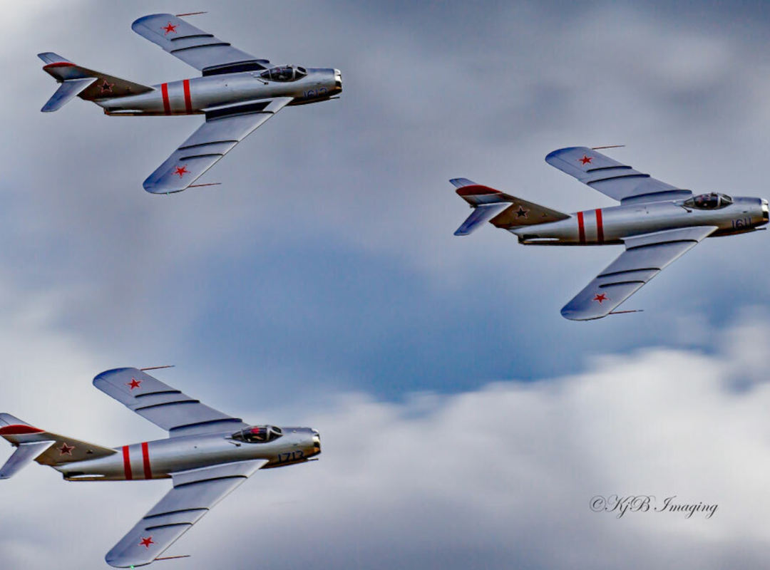 Three silver MiG fighter jets from the Fighterjets Jets Demo Team fly in formation against a cloudy sky. The aircraft feature red Soviet-style star markings and red fuselage stripes as they perform a coordinated aerial pass.