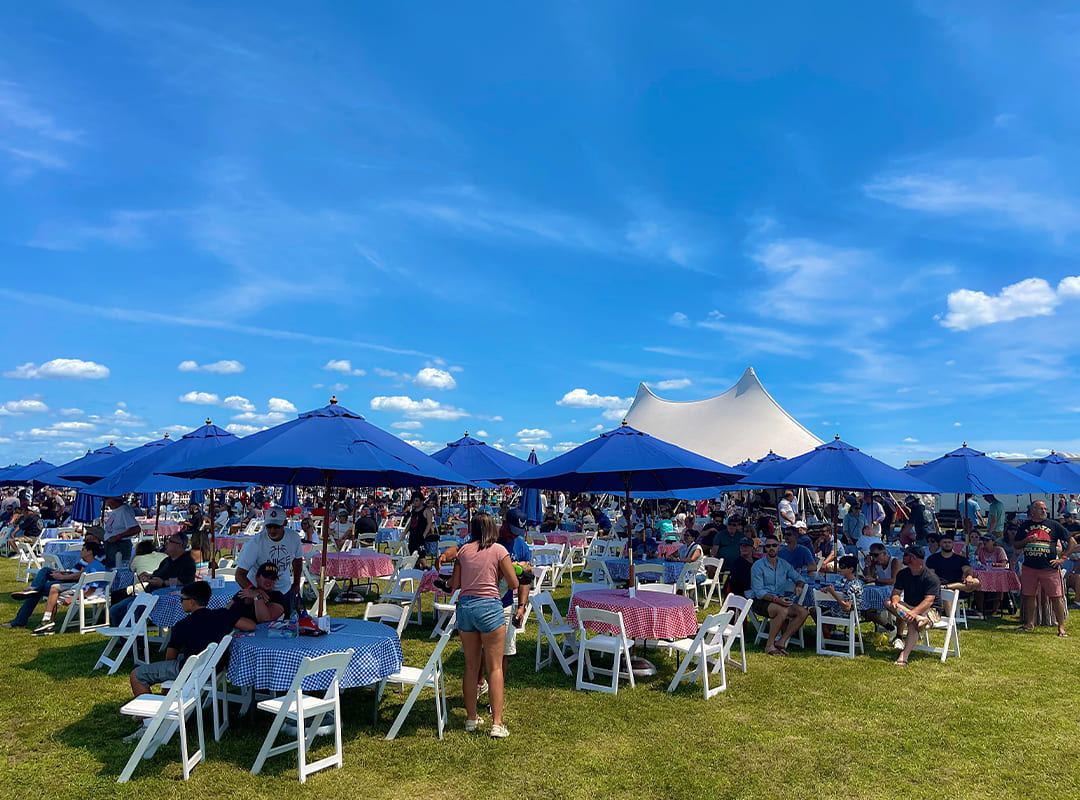 Outdoor air show hospitality area with guests seated at tables beneath blue umbrellas on a sunny day, with a large white tent and bright blue sky in the background.