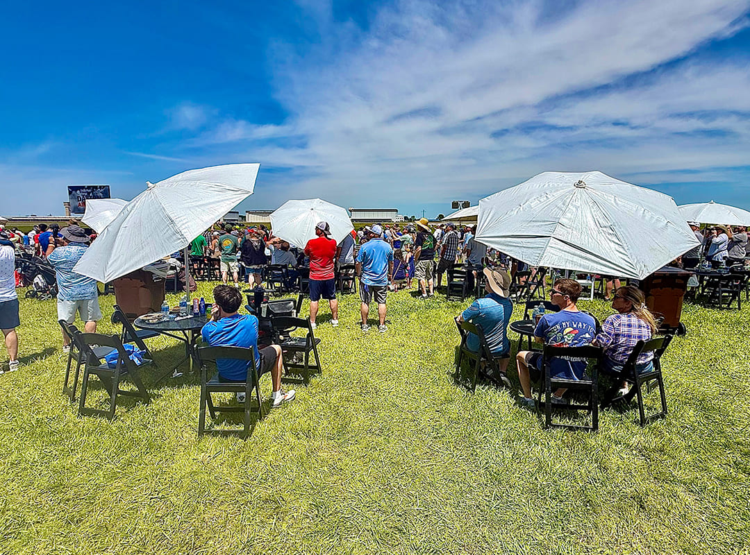 Air show guests seated at shaded tables and standing on a grassy viewing area beneath white umbrellas, watching the action under a bright blue sky.